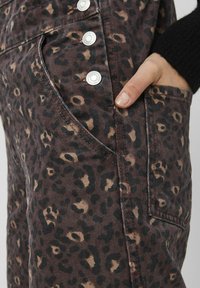 Tissu marron avec un motif léopard, équipé de ferrures métalliques argentées, poches latérales et finition texturée. Main placée dans la poche.