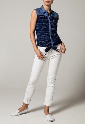 Chemise sans manches bleu marine à pois blancs, col de type denim, devant à boutons ; associée à un pantalon blanc ajusté et des chaussures blanches décontractées.