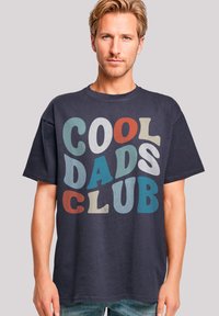 Navy blue cotton t-shirt featuring bold text "COOL DADS CLUB" in various colors (red, blue, gray, and cream) with a relaxed fit design.