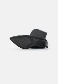 Bottines noires avec une tige en tricot texturé, bout pointu, semelle noire lisse et accent de talon. Équipées d'une languette pour un enfilage facile.