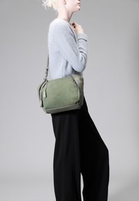 Borsa a spalla verde in suede con dettagli in pelle, dotata di chiusura con zip, dettaglio con nappina e tracolla regolabile. Indossata con un maglione grigio e pantaloni neri.