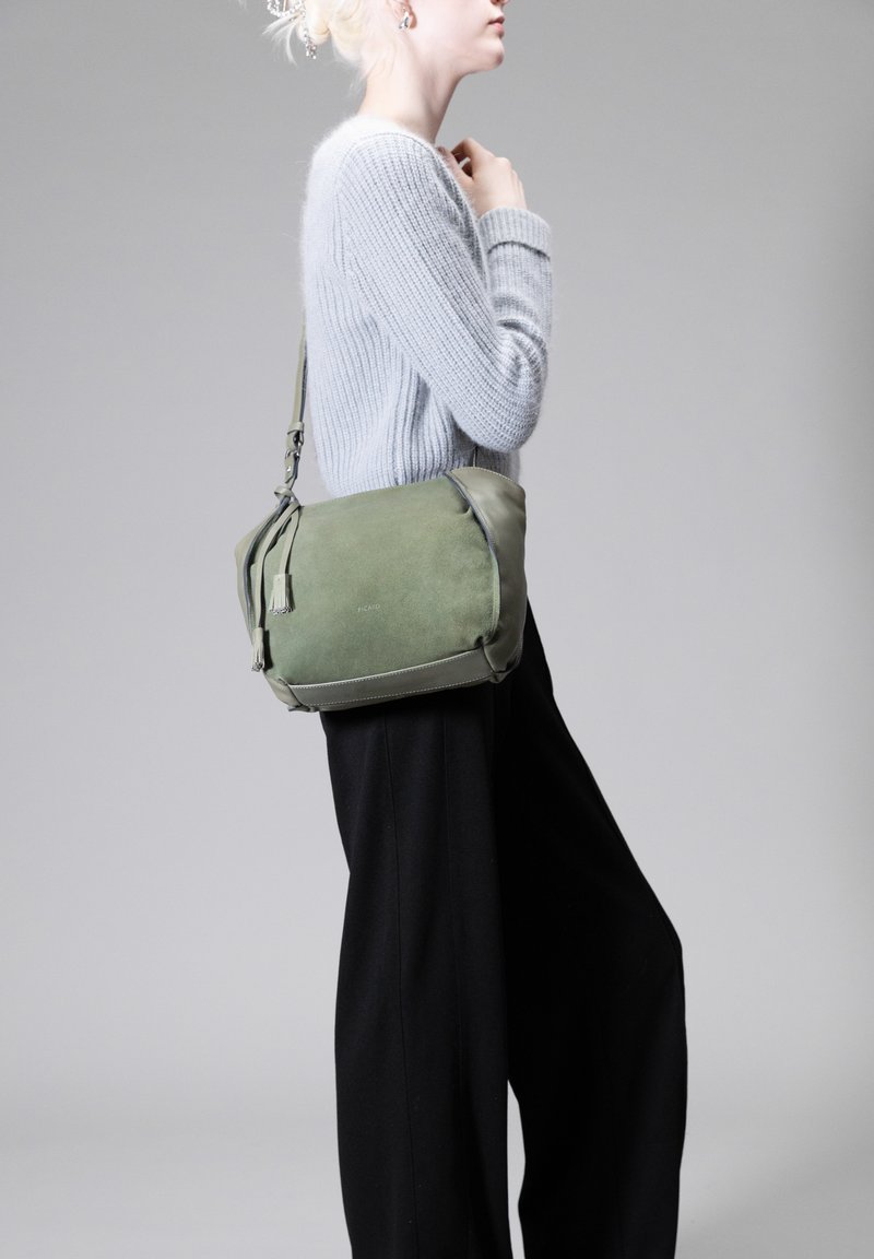 Borsa a spalla verde in suede con dettagli in pelle, dotata di chiusura con zip, dettaglio con nappina e tracolla regolabile. Indossata con un maglione grigio e pantaloni neri.