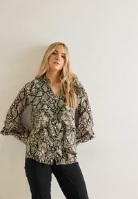 Leichte Bluse mit Schlangenprint in Schwarz, Creme und Oliv, V-Ausschnitt und rüschierten Glockenärmeln. Kombiniert mit schwarzen Hosen.