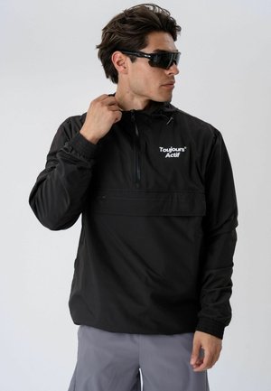 Mann trägt schwarze Zip-up-Jacke mit der Aufschrift "Toujours Actif" und dunkle Sonnenbrille, richtet den Kragen und steht vor hellem, einfarbigem Hintergrund.