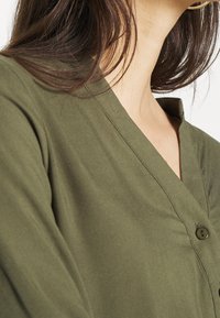 Blouse vert olive avec un décolleté en V, présentant un détail de bouton, une texture de tissu douce et une coupe ample, mettant en valeur l'épaule et la partie supérieure de la poitrine.