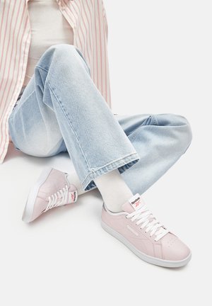 Persona sēž ar sakrustotām kājām, valkājot gaiši zilas džinsas, baltas zeķes un gaiši rozā Reebok sporta apavus ar baltām šņorēm.