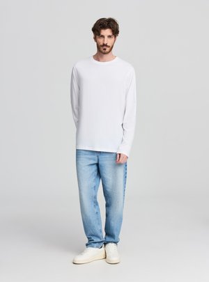 Maglietta bianca a maniche lunghe, materiale in cotone, scollo rotondo; abbinata a jeans taupe chiaro e scarpe da ginnastica bianche. Design semplice e casual.