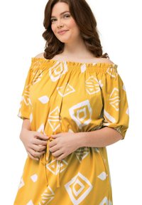 Keltainen olkapäältä avonainen mekko, jossa on valkoisia geometrisia kuvioita, lyhyet hihat, joustava kaula-aukko ja nyöri vyötäröllä. Sujuva puuvillakangas.