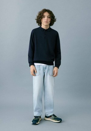 Polo a maniche lunghe blu navy, jeans a gamba dritta azzurri chiari e sneaker scure con dettagli verdi e beige, su sfondo grigio.