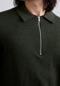 Polo de punto verde bosque con cremallera. Presenta un cuello y una tela texturizada. El detalle de la cremallera plateada añade contraste.
