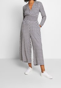 Mono de color azul marino con estampado, manga larga, escote en V profundo, cintura fruncida y piernas anchas y cortas; combinado con zapatillas blancas.