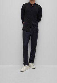 Camicia nera con bottoni e colletto alla coreana, abbinata a pantaloni scuri e sneakers bianche. La camicia ha maniche lunghe e una texture liscia.