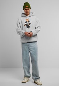 Grå hættetrøje med en bjørnegrafik, parret med lyseblå jeans og beige sneakers. En grøn hue med orange tekst fuldender outfittet.