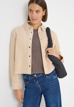 Beige corduroy overhemdjasje met drukknopen, twee borstzakken, gedragen over een bruine top, gecombineerd met blauwe jeans en een zwarte tas.