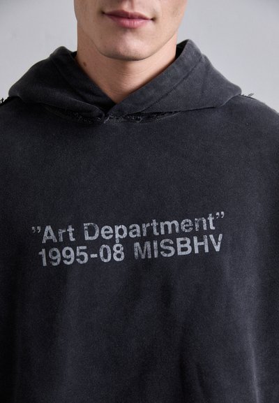 Junge Person trägt einen schwarzen Kapuzenpullover mit weißem Aufdruck "Art Department 1995-08 MISBHV" auf der Brust.