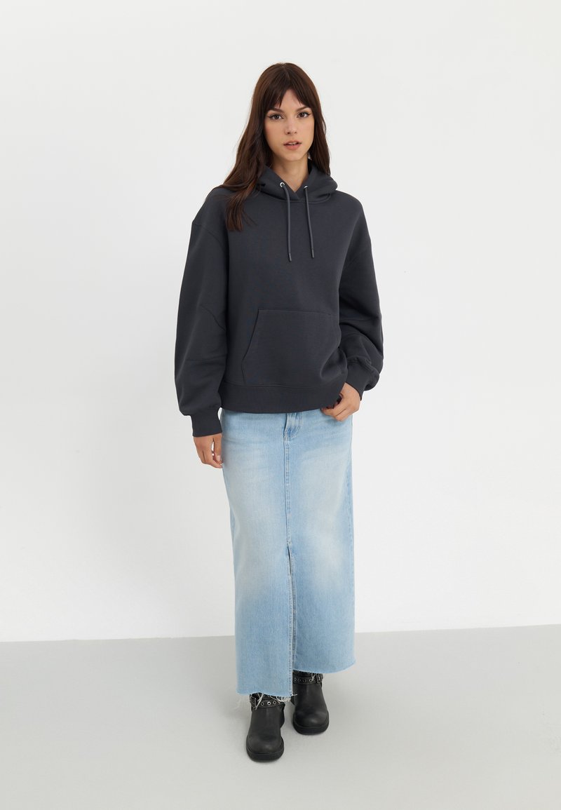 Szara bluza z kapturem z kieszenią z przodu, zestawiona z jasnoniebieską długą spódnicą dżinsową i czarnymi botkami na kostkę. Prosty, swobodny styl.