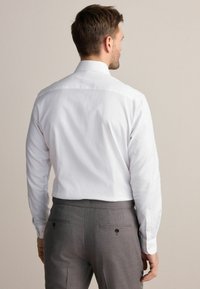 Muška bijela košulja s dugim rukavima, dugmadi na manžetama i klasičnim ovratnikom, u kombinaciji s sivim odijelom hlačama s detaljima na džepovima.