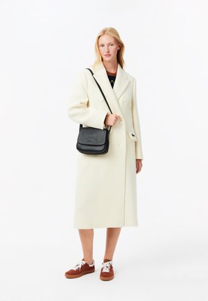 Cappotto crema doppiopetto con grandi bottoni, indossato sopra un maglione scuro, abbinato a una borsa a tracolla nera e sneakers marroni.