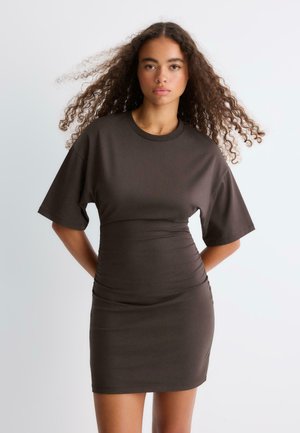 Femme aux cheveux bouclés portant une robe ajustée marron foncé à manches courtes, debout, les mains dans le dos, sur un fond clair uni.