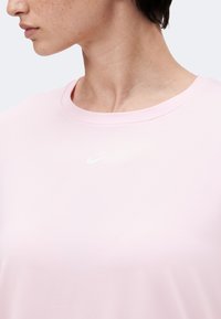 Γυναίκα που φοράει ανοιχτό ροζ μπλουζάκι Nike με στρογγυλή λαιμόκοψη, ορατή από τον λαιμό μέχρι το άνω μέρος του στήθους, κοιτάζει προς τα αριστερά.