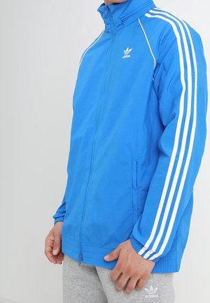Uomo che indossa una giacca da ginnastica Adidas blu con strisce bianche sulle maniche e pantaloni della tuta Adidas grigi, in piedi davanti a uno sfondo semplice.