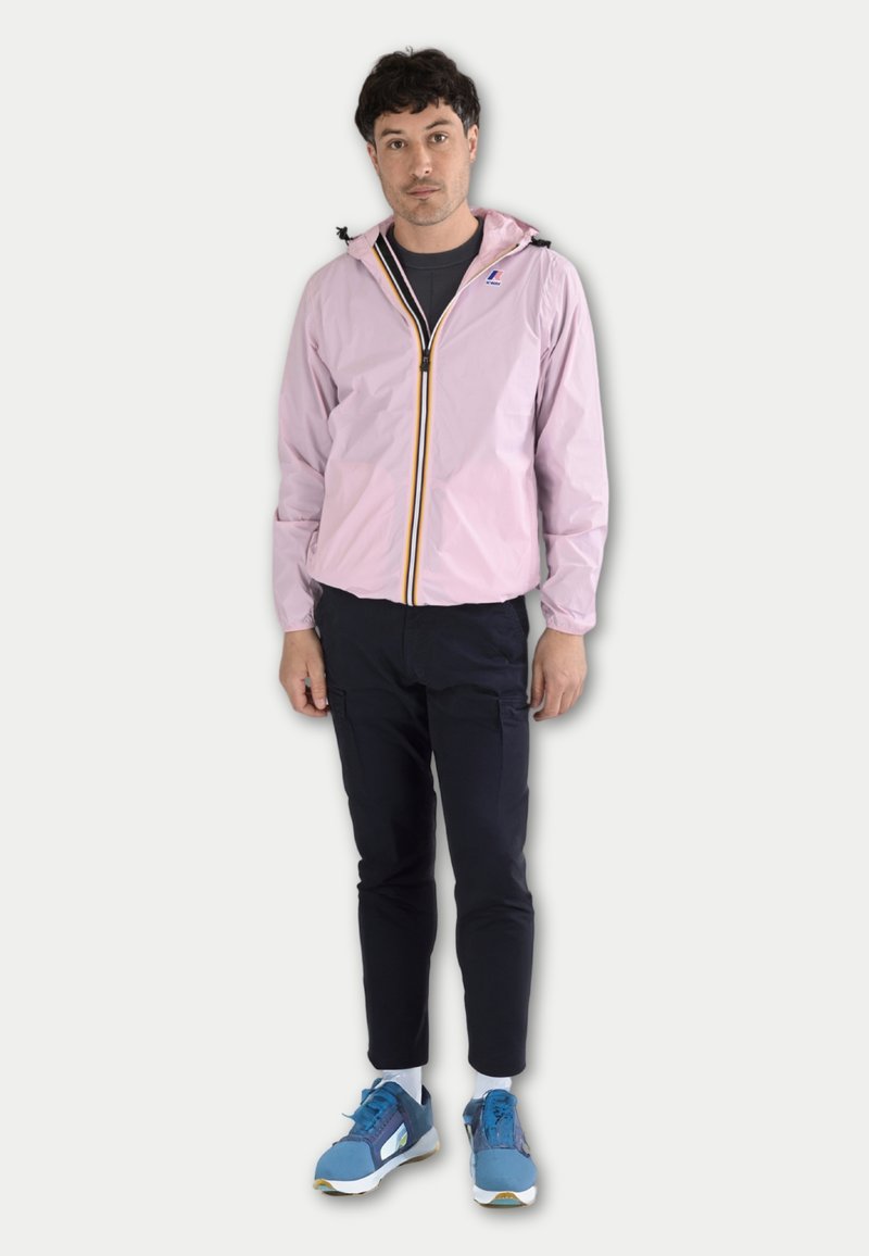 Coupe-vent rose clair avec capuche, fermeture éclair noire et accent jaune. Porté sur une chemise sombre, associé à un pantalon bleu marine et des chaussures bleues.