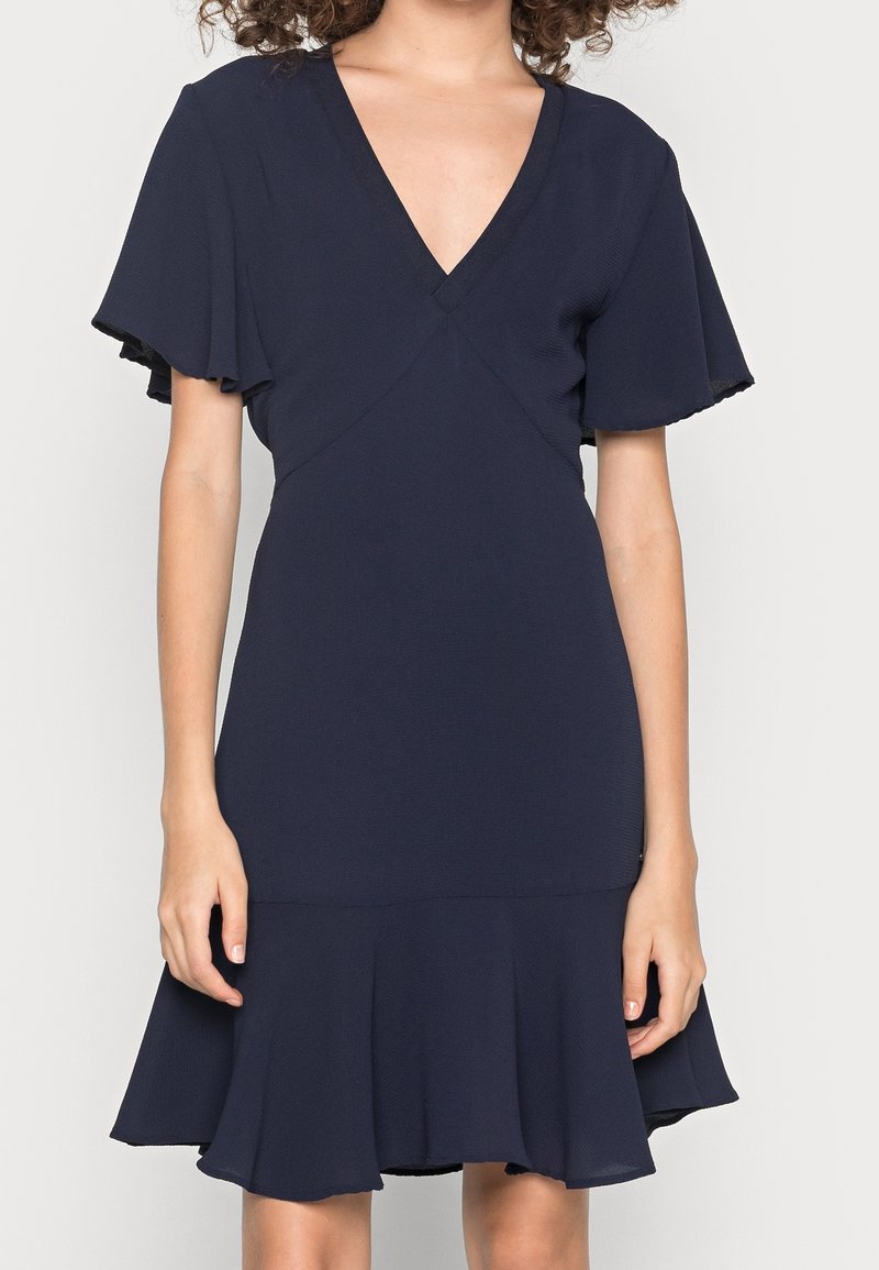 Femme portant une robe bleu marine foncé avec des manches courtes volantées, un décolleté en V, une taille cintrée et un ourlet à volants évasé.