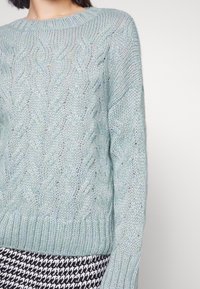 Ljusgrön stickad tröja med kabelmönster, rund halsringning och textilstruktur; bärs över ett plagg med svartvit houndstooth-mönster.