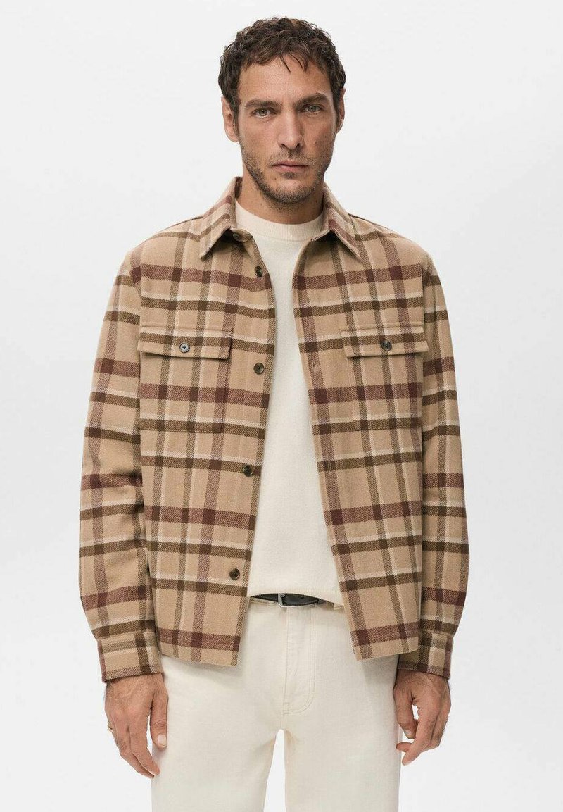 Chaqueta de cuadros marrón y tostado en tela suave, con dos bolsillos en el pecho, botones delanteros y un diseño clásico de cuello. Lleva puesta una camisa color crema debajo.