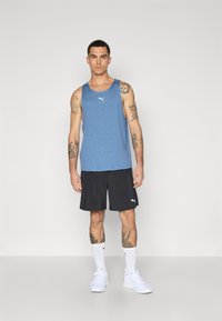 Blå atletisk tanktop med et hvidt logo, parret med sorte shorts og hvide sneakers. Modellen har synlige tatoveringer og hvide sokker.