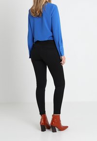 Kvinna klädd i en blå blus, svarta slim fit byxor och bruna klackade ankelboots som står mot en enkel vit bakgrund.