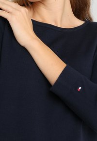 Námorně modrý dlouhý rukáv top vyrobený z žebrované tkaniny. Má lodičkový výstřih a malý akcent loga v červené, bílé a modré barvě na rukávu.