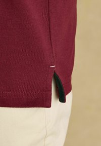 Küljelt vaade inimesest, kes kannab burgundia värvi tekstuuriga lühikeste varrukatega särki, millel on väike külgauk, heledate beežide pükste peal.