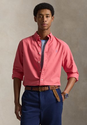 Férfi korall színű, gombos ingben csíkos póló fölött, tengerészkék nadrágban, fonott barna övvel és karórával, semleges háttér előtt pózol.