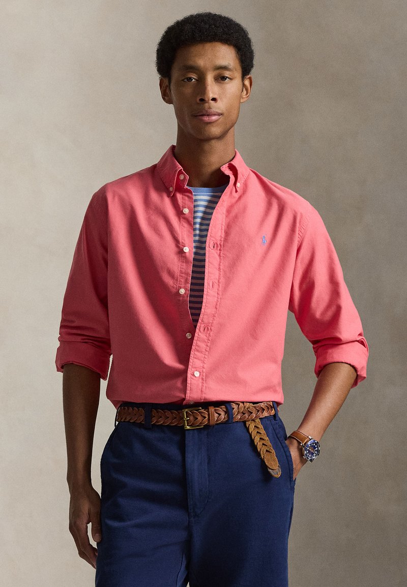 Muž nosící korálovou košili na knoflíky přes pruhované tričko, tmavě modré kalhoty s pleteným hnědým páskem a náramkové hodinky, pózující na neutrálním pozadí.