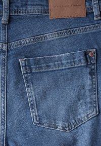 Primer plano de un bolsillo trasero de unos jeans de mezclilla azul con una etiqueta de cuero marrón que dice "Street One Studio."