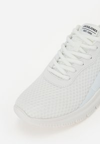 Λευκό αθλητικό παπούτσι με επάνω μέρος από δίχτυ, τόνοι κορδονιών και υφή στη σόλα. Περιλαμβάνει μια μικρή ετικέτα λογότυπου στη γλώσσα και λεία δερμάτινα στοιχεία.