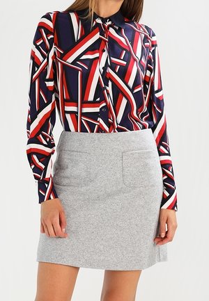 Kobieta ubrana w bluzkę z długim rękawem o geometrycznym wzorze w kolorach czerwonym, granatowym i białym oraz jasnoszarą spódnicę do kolan z przednimi kieszeniami.
