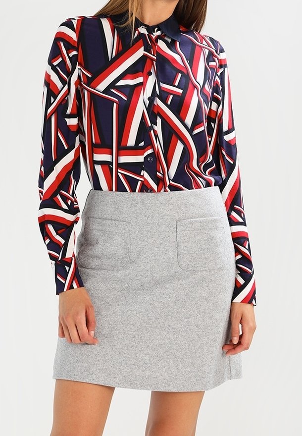 Femme portant un chemisier à manches longues à motif géométrique rouge, marine et blanc, avec une jupe grise claire mi-longue à poches avant.