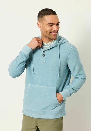 Mann lächelt, trägt ein hellblaues Kapuzensweatshirt mit Knöpfen, Hand in der Tasche, steht vor einem einfarbigen hellen Hintergrund.