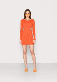 Vestido laranja de manga longa com tecido texturizado, recortes na cintura, silhueta justa, combinado com sapatos de salto laranja e óculos de sol.