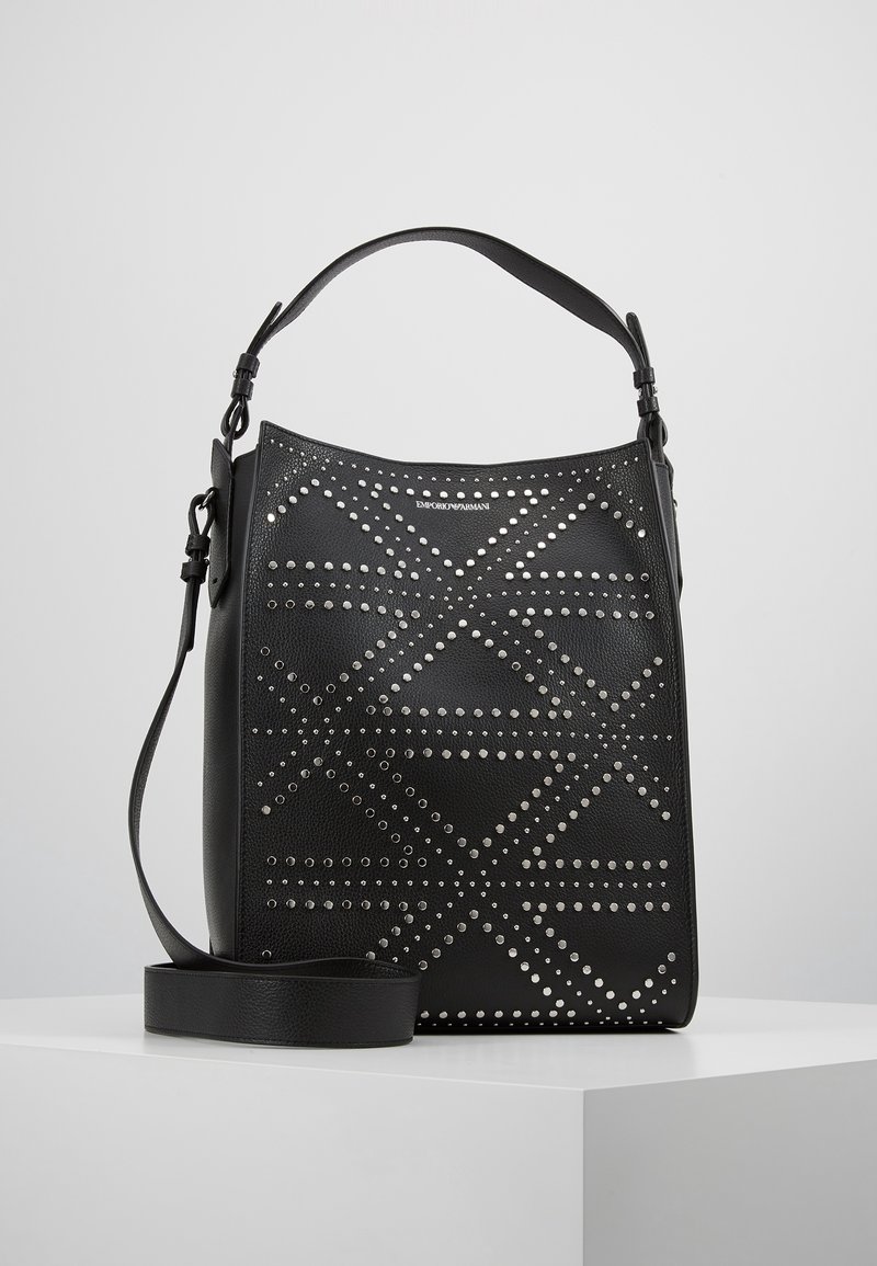 Czarna skórzana torba z perforowanymi geometrycznymi wzorami, ozdobiona srebrnymi ćwiekami, z płaskim paskiem na ramię i wyraźnie zarysowanym kształtem.