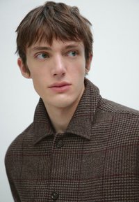 Brun ternet jakke med krave, der har store knapper, vævet tekstur og subtile røde accenter. Skræddersyet pasform, afslappet design.