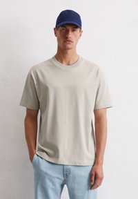 Beige T-shirt met korte mouwen en een ronde hals, gecombineerd met lichtblauwe jeans. De model draagt een marineblauwe pet en heeft de handen in de zakken.