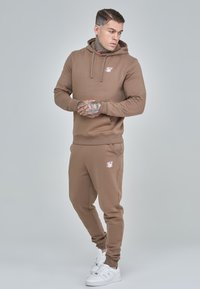 Brown cotton hoodie with a front pocket and trim, paired with matching joggers featuring an emblem and elastic cuffs. White sneakers accent the outfit.