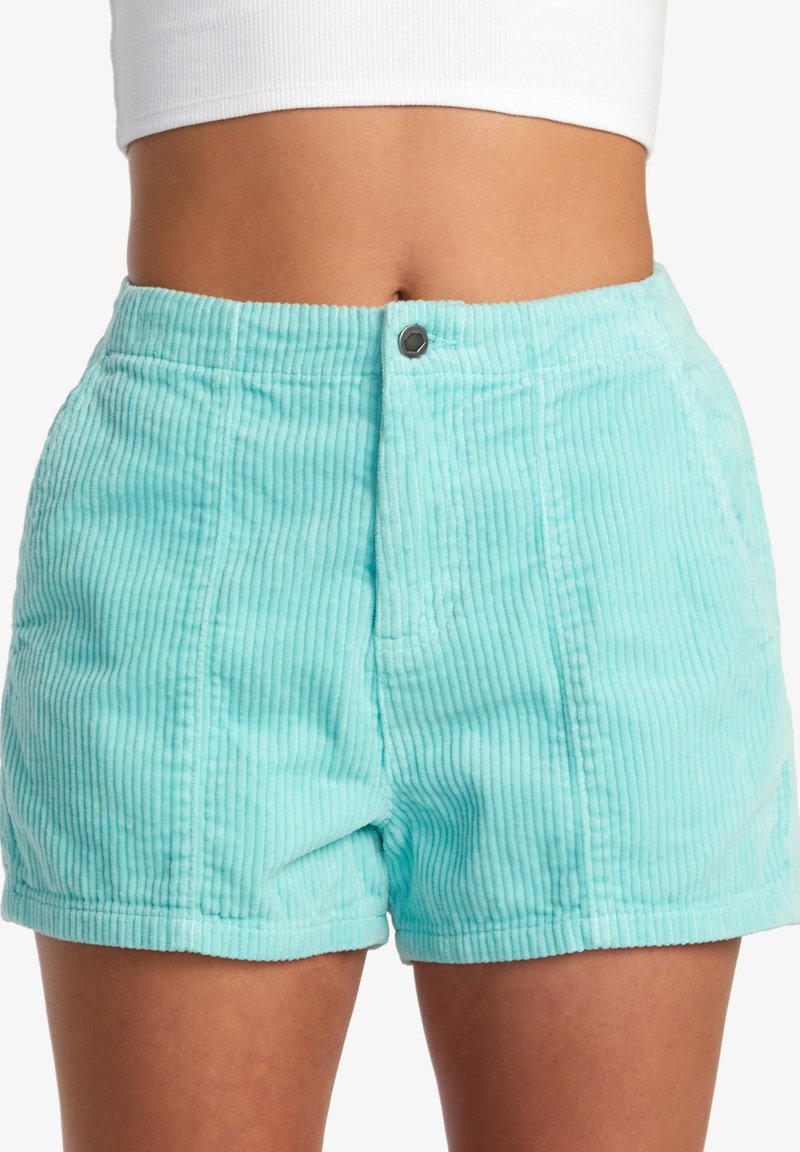 Donna che indossa pantaloni corti in corda di colore turchese chiaro a vita alta e un top bianco corto, mostrata dalla vita a metà coscia.