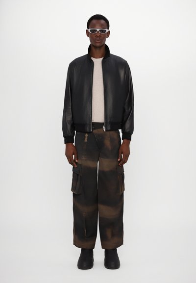 Zwarte leren bomberjack, grijze trui, oversized bruine cargobroek met zakken en zwarte laarzen. Accessoires zijn witte zonnebril.
