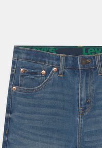 Blaue Denim-Jeans mit Kupferriegeln, orangefarbener Naht, Knopfverschluss und sichtbarem grünen Levi's-Logo am Bund.