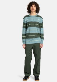 Langarmgestreiftes T-Shirt in Hellblau, Dunkelgrün und Gelb. Kombiniert mit grüner Hose und schwarzen Turnschuhen. Verfügt über eine Brusttasche.