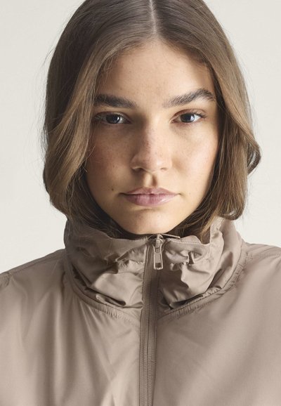 Junge Frau mit hellbraunen Haaren, die eine beige Jacke mit Reißverschluss trägt, schaut mit neutralem Gesichtsausdruck nach vorne vor einem schlichten Hintergrund.